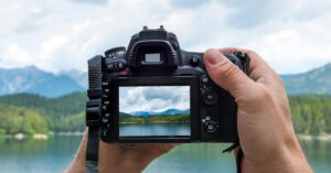 Hands of a male photographer holding a digital camera taking pictures of a idyllic landscape with a lake and mountains while the picture shows at the display