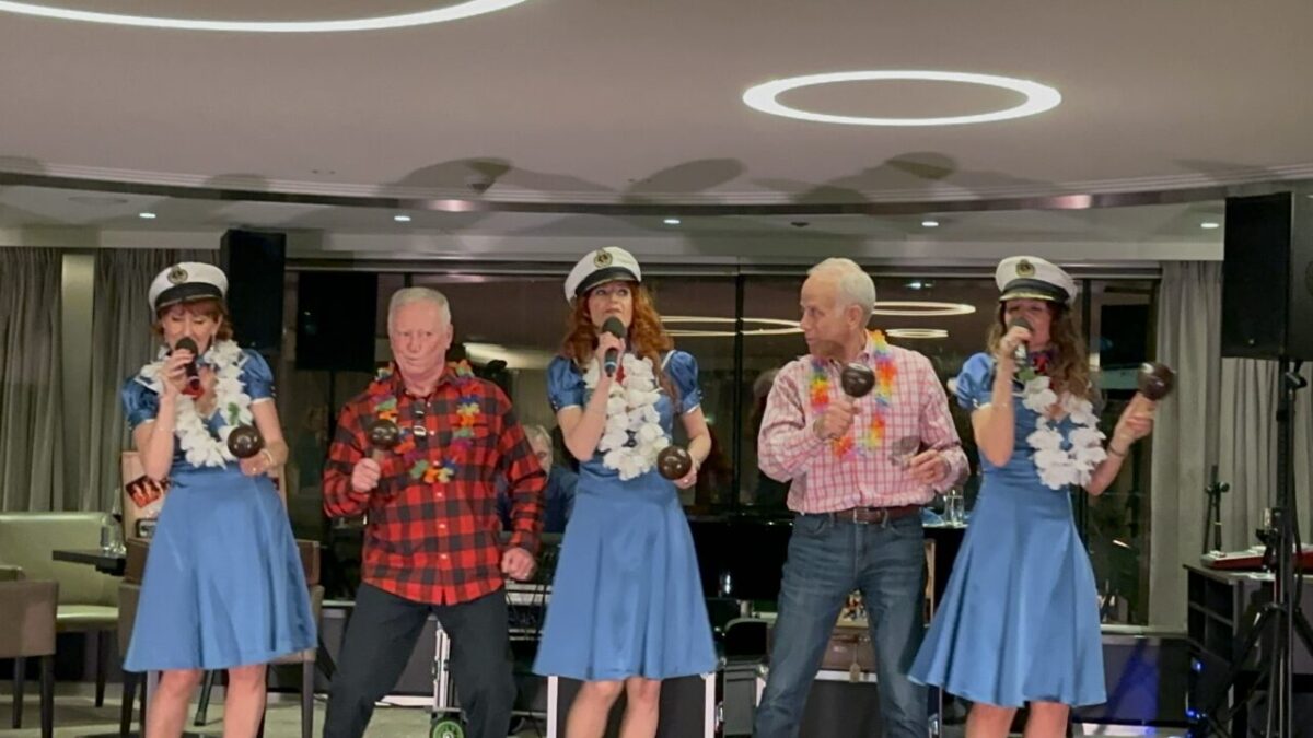 Ship crew and passengers sing a song on stage.